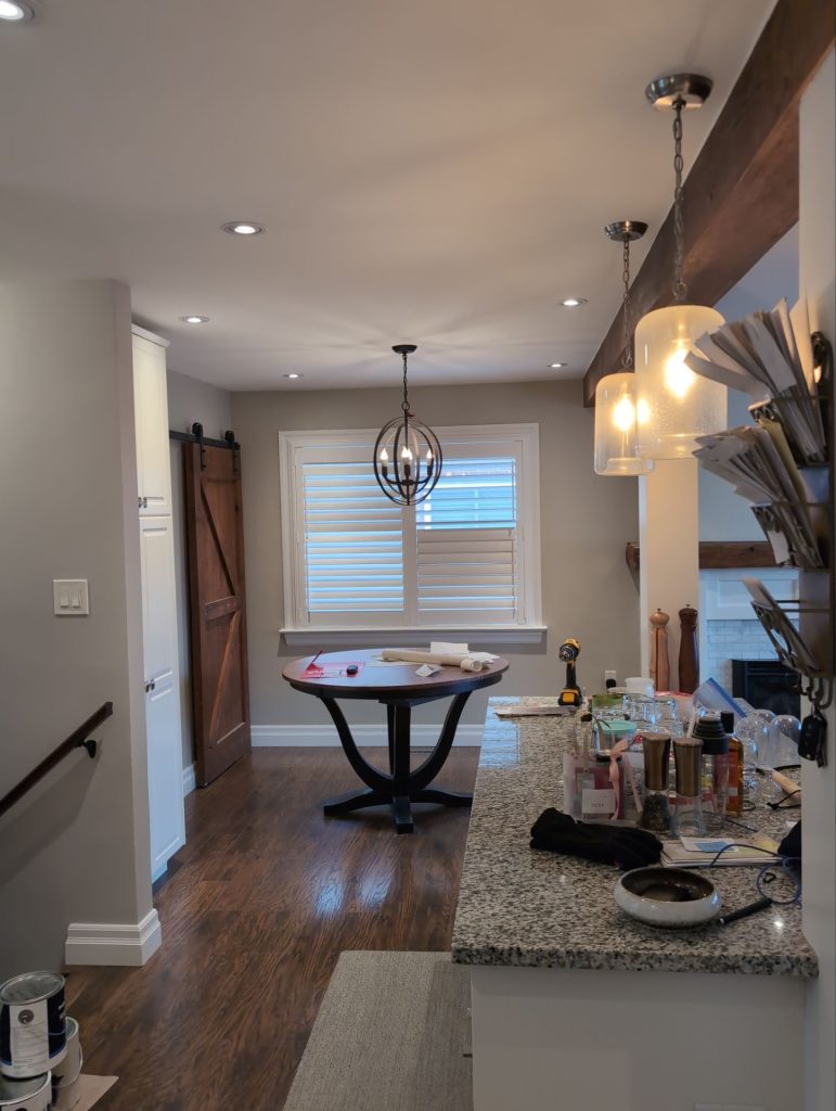 A bright kitchen and dining area with a round table, pendant lighting, and granite countertop, featuring warm wood floors, a sliding barn door, and natural light through plantation shutters.
